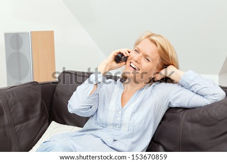 A beautiful blond woman is sitting on a couch, talking into the phone, having a conversation. She is laughing.