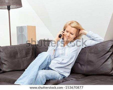 A beautiful blond woman is sitting on a couch, talking into the phone, having a conversation.