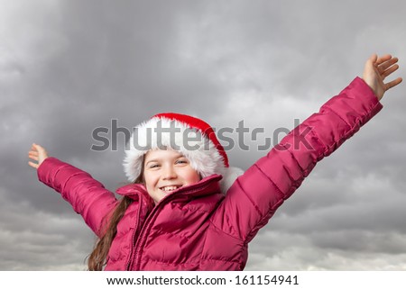 Cute young girl standing outside, wearing a red Santa hat,looking into the camera, her arms stretched out up in the air