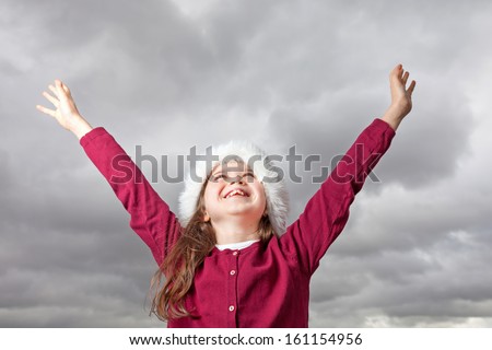 Cute young girl standing outside, wearing a red Santa hat, her arms stretched out up in the air