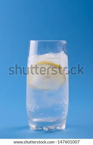 Summer fresh drink - cold sparkling water in a glass with some ice and slice of a lemon over a solid blue background.
