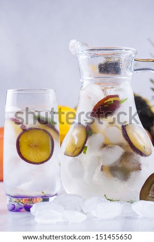 Fresh fruit and water drink with a purple plums slices, mint herb and ice cubes with some fruits in the background.