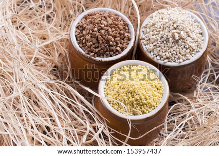 Close up photo of a eco raw food in the clay cup - light brown pearl barley, dark brown kasha and yellow millet on a  wooden shavings.