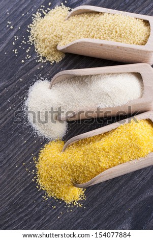 Close up photo of a raw eco food in the wooden spatulas - light yellow couscous, light brown farina and dark yellow cornmeal placed on a dark wooden background.