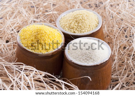 Close up photo of a raw eco food in the clay cup - light yellow couscous, light brown farina and dark yellow cornmeal placed on a dark wooden background.
