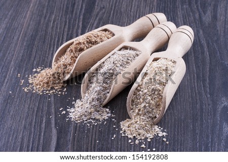 Close up photo of a cereal grain product in a wooden scoop - dark brown wheat bran, light brown secale bran, and mid-brown arena bran placed on a dark wooden background.