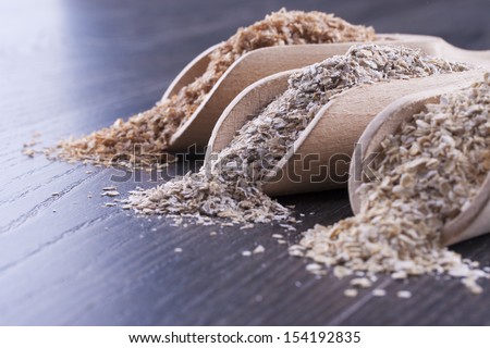 Close up photo of a cereal grain product in a wooden scoop - dark brown wheat bran, light brown secale bran, and mid-brown arena bran placed on a dark wooden background.