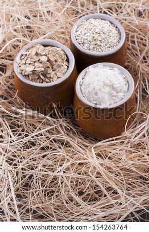Close up photo of a cereal grain product in a clay cup - light brown barley flakes, white rice flakes and dark brown secale flakes placed on a wooden shavings.