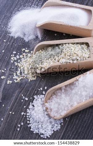 Close up photo of a food ingredients in a wooden scoop - small pieces of a table salt, yellow colored garlic salt and large pieces of a sea salt placed on a dark wooden background.