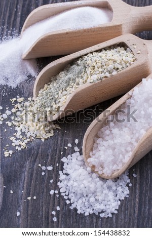 Close up photo of a food ingredients in a wooden scoop - small pieces of a table salt, yellow colored garlic salt and large pieces of a sea salt placed on a dark wooden background.