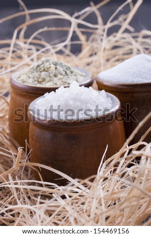 Close up photo of a food ingredients in a clay cup - small pieces of a table salt, yellow colored garlic salt and large pieces of a sea salt placed on a wooden shavings.