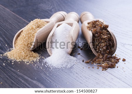 Close up photo of a food ingredients in a wooden scoop - brown sugar cane, white sugar and large pieces of crystalized sugar placed on a dark wooden background.