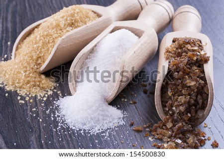Close up photo of a food ingredients in a wooden scoop - brown sugar cane, white sugar and large pieces of crystalized sugar placed on a dark wooden background.