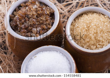 Close up photo of a food ingredients in a clay cup - brown sugar cane, white sugar and large pieces of crystalized sugar placed on a wooden shavings.