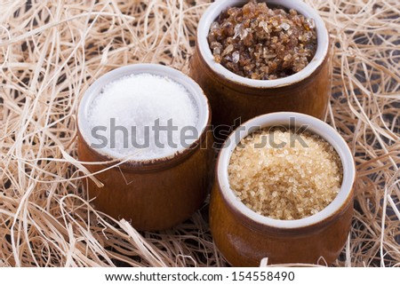 Close up photo of a food ingredients in a clay cup - brown sugar cane, white sugar and large pieces of crystalized sugar placed on a wooden shavings.