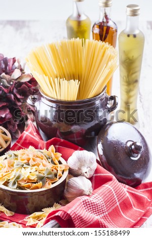 Raw food composition - brown penne, yellow, orange and green farfalle and yellow spaghetti  pasta in a clay pot placed on a bright wooden background.