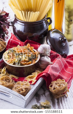 Raw food composition - brown penne, yellow, orange and green farfalle, brown tagliatelle and yellow spaghetti  pasta in a clay pot placed on a bright wooden background.