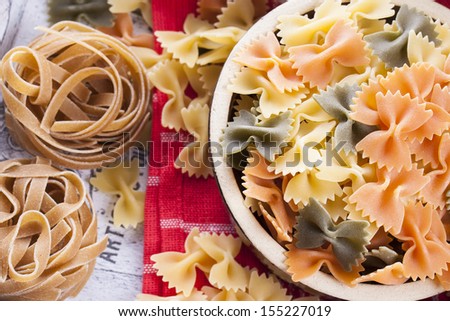 Raw food composition - yellow, orange and green farfalle, brown tagliatelle in a clay pot placed on a bright wooden background.