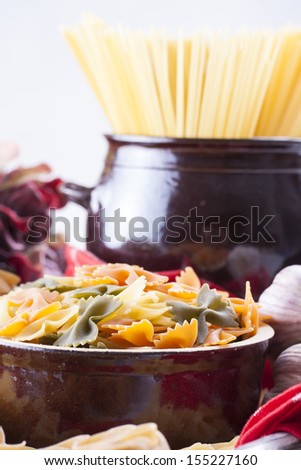 Raw food composition - yellow, orange and green farfalle and yellow spaghetti  pasta in a clay pot placed on a bright wooden background.