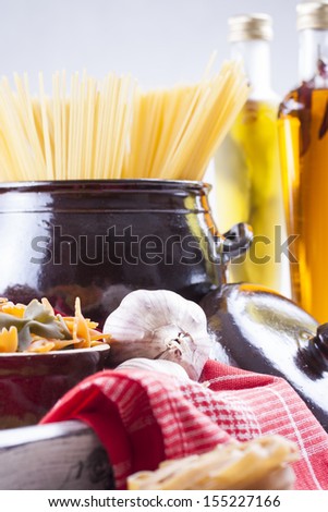 Raw food composition - brown penne, yellow, orange and green farfalle and yellow spaghetti  pasta in a clay pot placed on a bright wooden background.