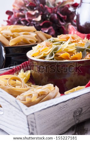 Raw food composition - brown penne, yellow, orange and green farfalle, brown tagliatelle pasta in a clay pot placed on a bright wooden background.