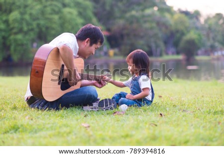 Asian father is playing guitar nearly with his daughter sitting and touching the guitar in the background of green park with happiness moment, concept of activity in family lifestyle.