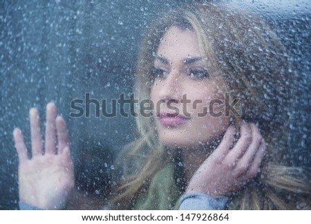 Thoughtful young woman looking through glass window with raindrops