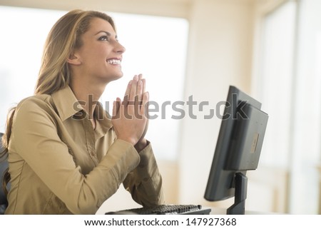 Happy young businesswoman with hands clasped praying while sitting at desk in office