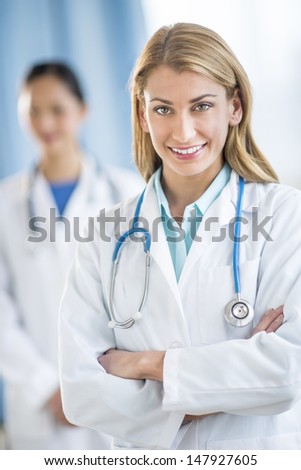 Portrait of happy young female doctor standing arms crossed at clinic with colleague in background