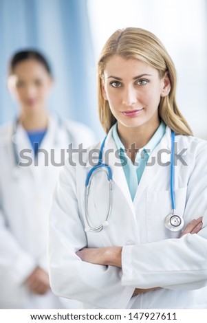 Portrait of confident female doctor standing arms crossed with colleague in background at clinic