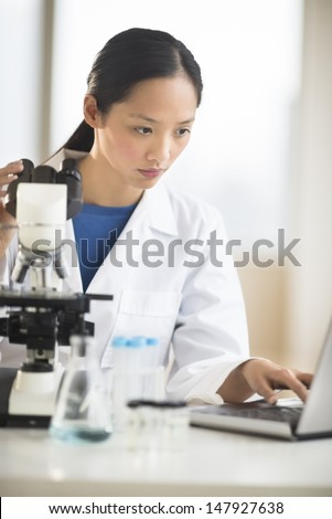 Mid adult Asian female doctor using laptop with microscope at desk in laboratory