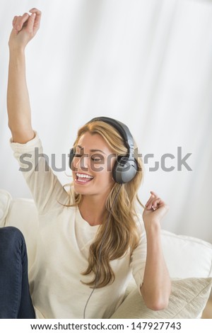 Happy young woman with hand raised enjoying music through headphones while sitting on sofa at home