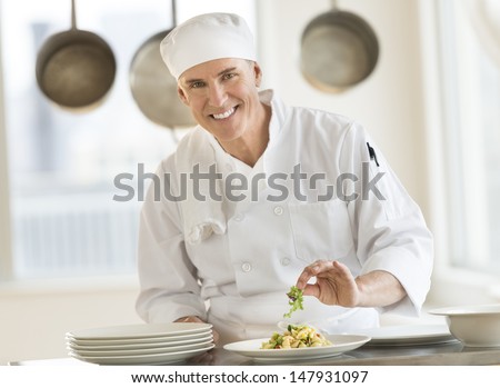 Portrait of happy mature male chef garnishing pasta dish at counter in commercial kitchen