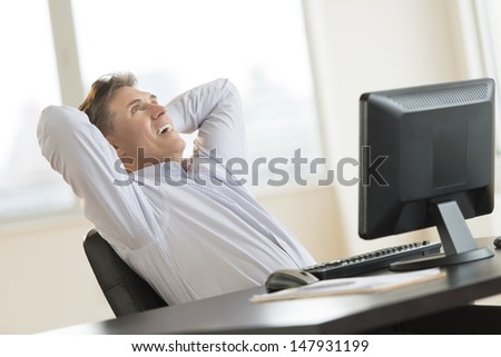 Happy mature businessman with hands behind head looking up while sitting at desk in office
