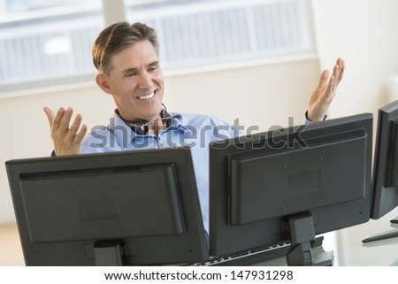 Happy mature male trader gesturing while using multiple screens at desk in office