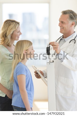 Mature male doctor measuring girl's weight while looking at woman in hospital