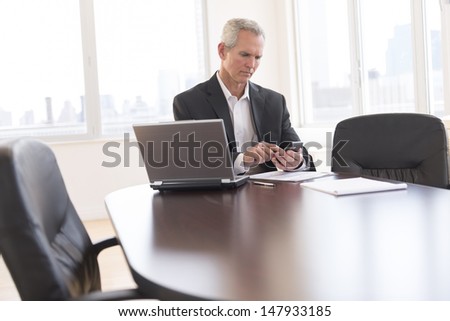 Mature businessman touching smart phone while sitting at desk in office