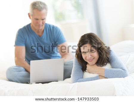 Portrait of happy mature woman relaxing in bed while man using laptop at home