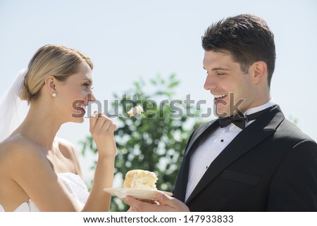 Happy young bride feeding wedding cake to groom outdoors
