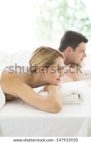 Side view of beautiful young woman resting on massage table with man in background at health spa