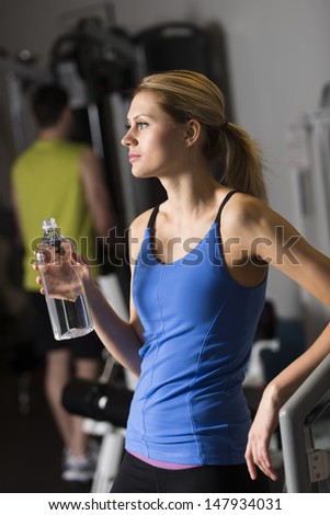 Thoughtful young woman with water bottle looking away while man standing in background at gym