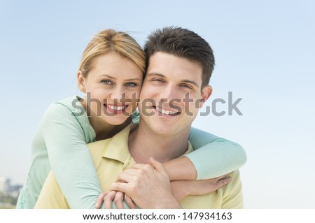 Closeup portrait of beautiful young woman embracing man from behind against clear blue sky