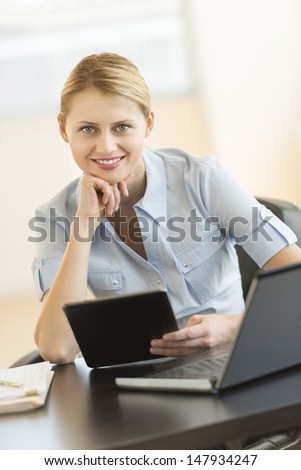 Portrait of confident young businesswoman with hand on chin holding digital tablet at office desk