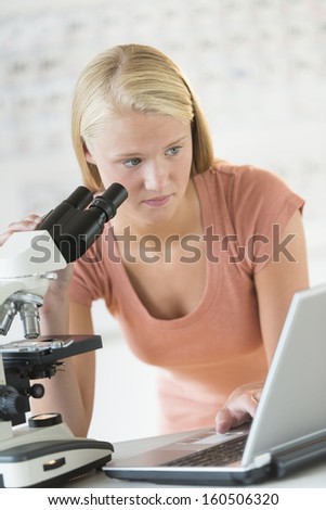 Teenage student looking at laptop while using microscope in chemistry class