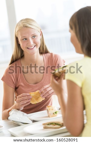 Happy teenage girl having snacks with friend in university canteen
