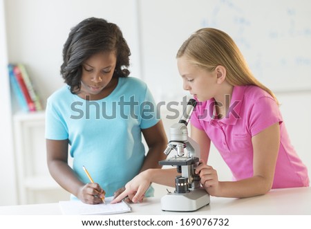 Multi-ethnic female students with microscope making notes in science class