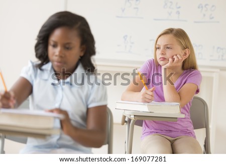 Thoughtful schoolgirl with hand on chin looking away while student writing notes in classroom
