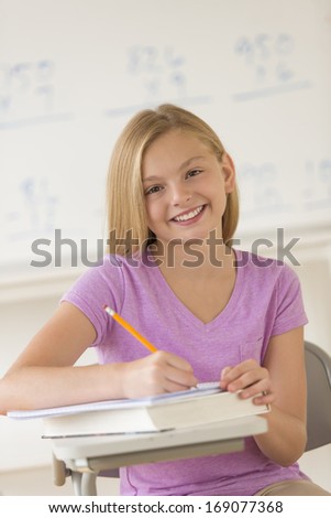 Portrait of happy girl writing notes in book at classroom desk