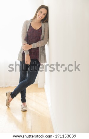Full length portrait of smiling young woman leaning on wall at home