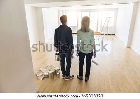 Full length rear view of mature couple with paint equipment standing on hardwood floor at home